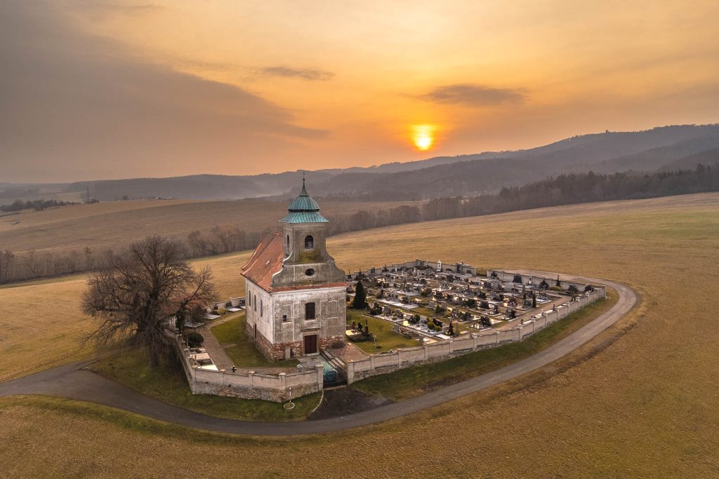 Pohled na historický kostel sv. Jakuba s malým hřbitovem obklopeným zdí, stojícím osamocen uprostřed pole poblíž silnice, při východu slunce