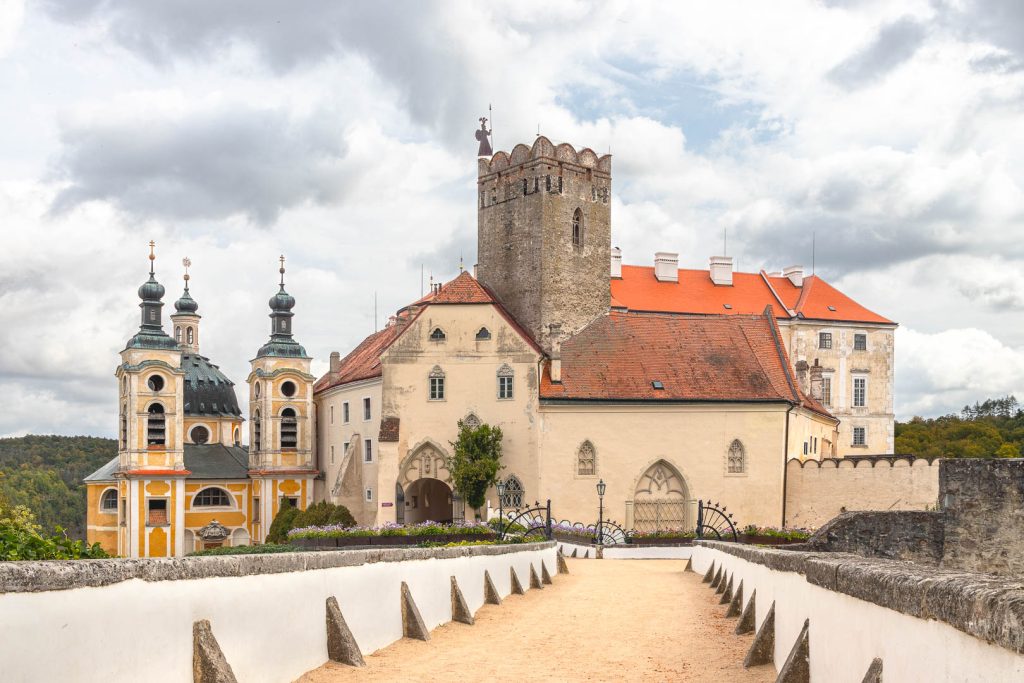 Pohled z mostu na zámek Vranov nad Dyjí a zámeckou kapli, která je hned vedle něj. Most je pokrytý pískem, ve stejné barvě je i fasáda zámku, má červenou střechu a vysokou věž. Kaple je žlutobílá se zelenou střechou. Je zataženo. Dramatické mraky.