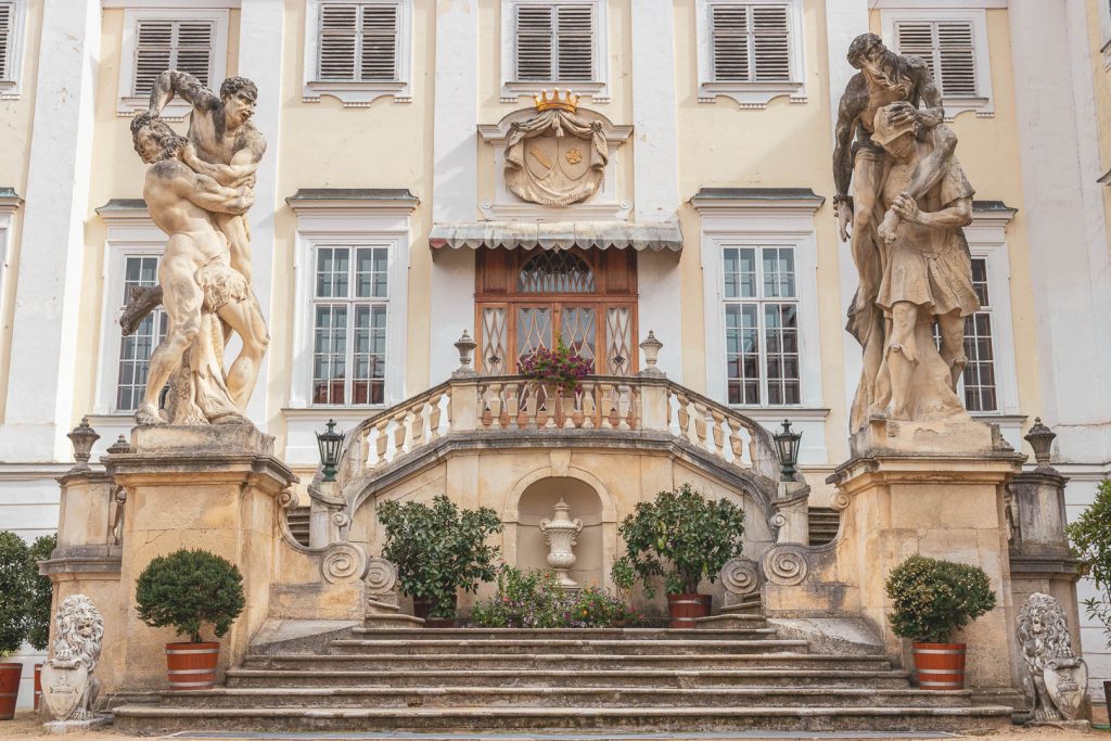 Čelní pohled na schody nádvoří vedoucí do sálu zámku Vranov nad Dyjí, schody vedou mezi dvěma velkými sochami, jsou pokryté květinami, měkké světlo tvoří jemné stíny na pískové omítce.