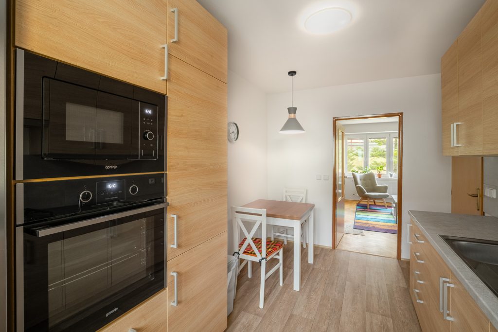 Moderní minimalistická kuchyně - pohled směrem k obývacímu pokoji, vestavěné spotřebiče (mikrovlnka, trouba), jídelní stůl s židlemi.