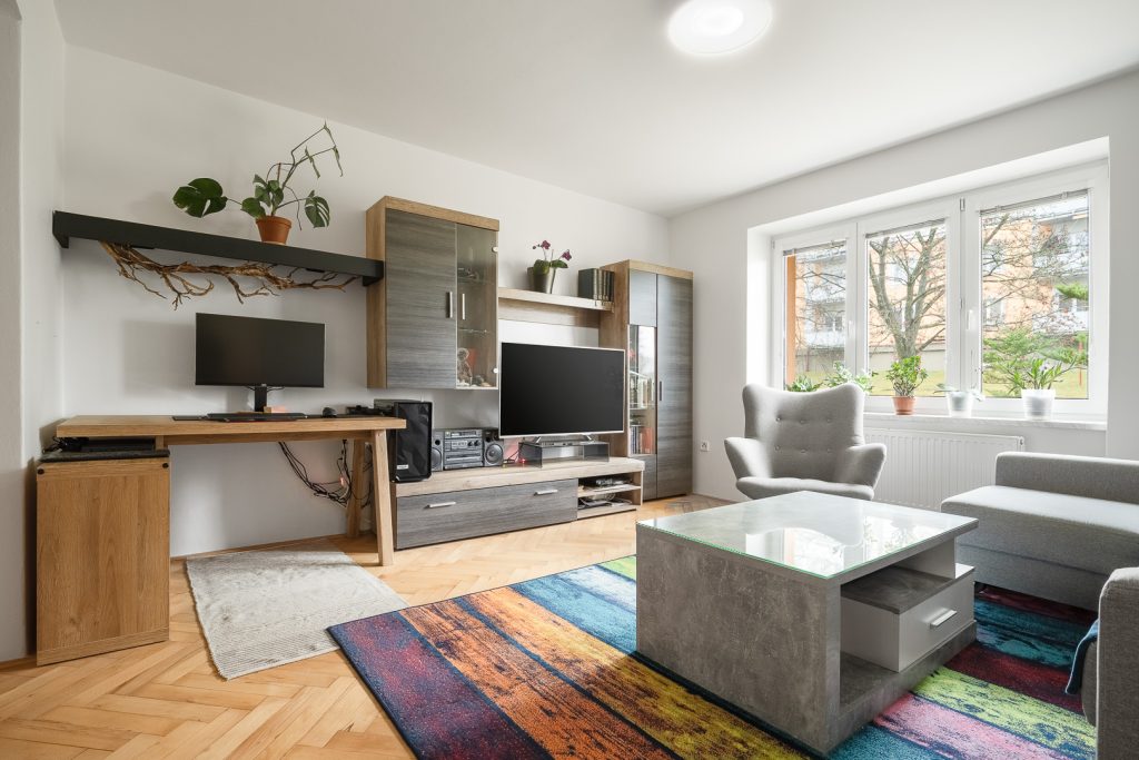 Prosvětlený minimalistický moderní obývací pokoj s parketami, šedým konferenčním stolkem a sedačkou, dřevěným nábytkem, barevným žíhaným kobercem a bílými stěnami.