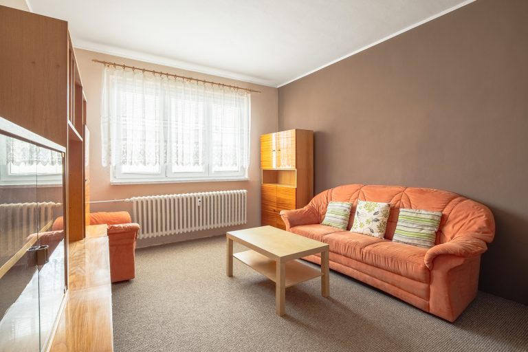 Prosvětlený obývací pokoj se světle hnědým dřevěným nábytkem, hnědým kobercem a hnědými stěnami, bílý strop, oranžová sedačka a křeslo, velké okno